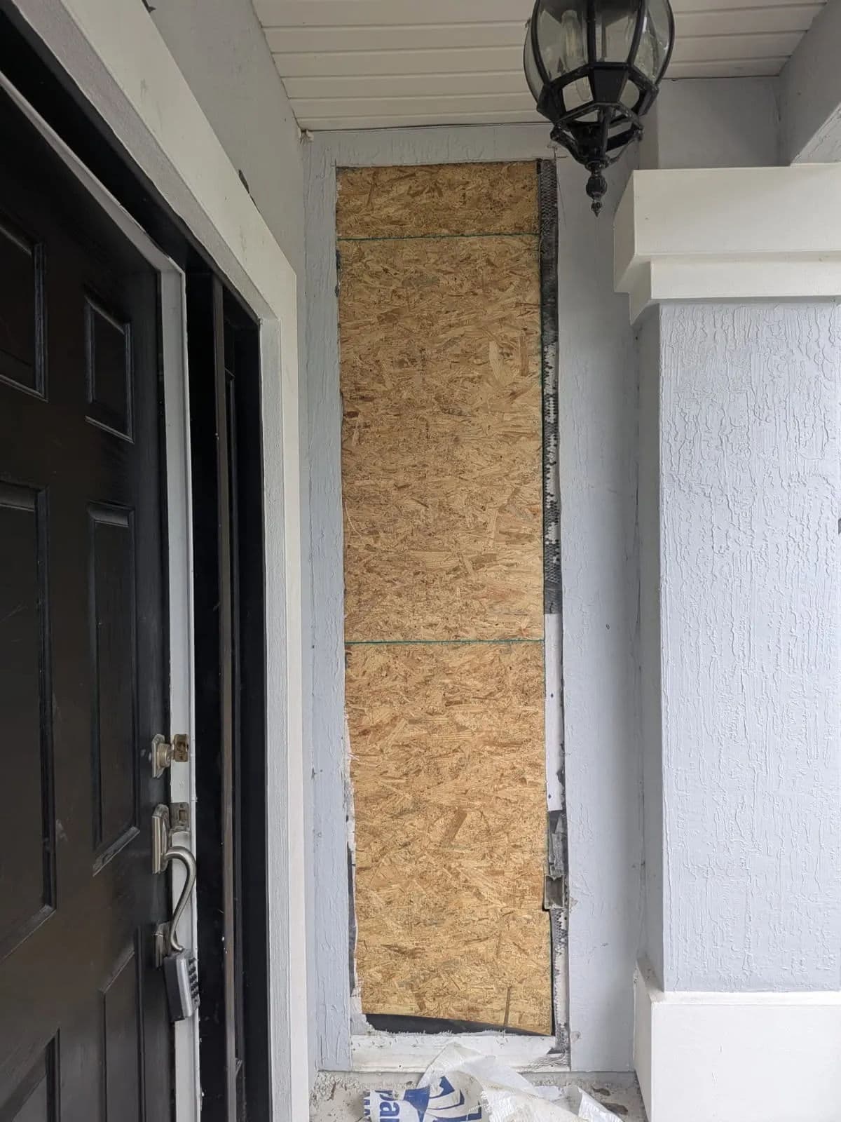 Boarded-up doorway with OSB plywood, near a black front door and a wall-mounted lantern.